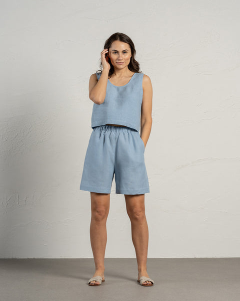 Een vrouw met donker haar draagt een korte broek van 100% linnen in de kleur wolkachtig blauw en een linnen top in dezelfde kleur. Haar ene hand zit in de zak terwijl de andere haar haar aanraakt. De achtergrond van de foto is in natuurlijke, neutrale tinten.