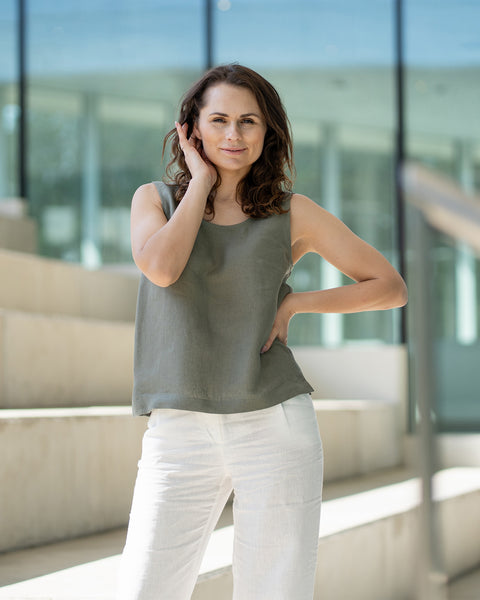 A woman with brown hair, wearing a stone green sleeveless linen top and white trousers, stands on wide concrete stairs inside a modern building with glass walls.
