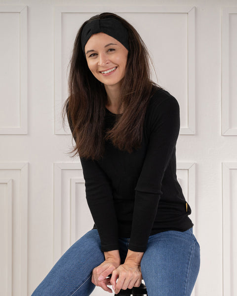 A smiling woman sitting and looking at the camera, wearing a black knit bow headband, a black long-sleeved top, and blue jeans.