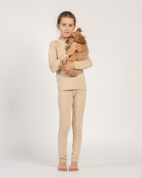 Meisje draagt een beige bamboe shirt met lange mouwen en een broek, staat blootsvoets en houdt een kleine bruine poedel in haar armen vast.
