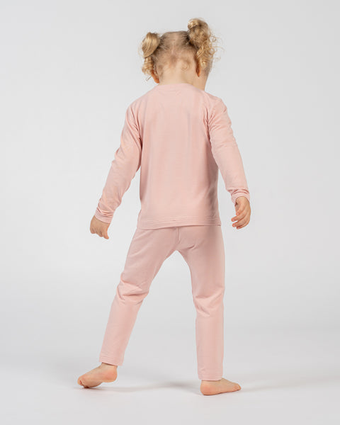 Kind in een roze shirt met lange mouwen en bijpassende broek, staat blootsvoets met de rug naar de camera en de armen ontspannen langs het lichaam, toont de kleding van achteren tegen een effen achtergrond.