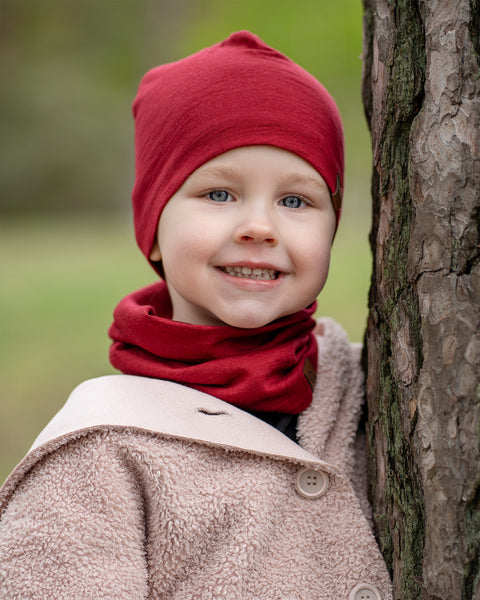 A young child with light skin and blue eyes is smiling broadly while leaning against the trunk of a tree. They are wearing a menique royal cherry beanie, a matching neck gaiter, and a light-colored, possibly beige, textured coat.