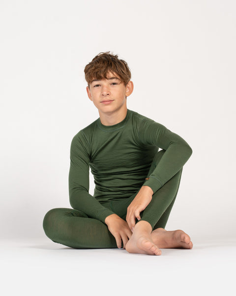 A barefoot child sits comfortably on the floor against a plain white background, wearing a matching dark green long-sleeve top and leggings. One leg is bent with their arm resting on their knee, while they look calmly at the camera.
