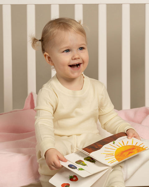 Happy toddler sitting in the crib, holding a book and wearing merino wool set.