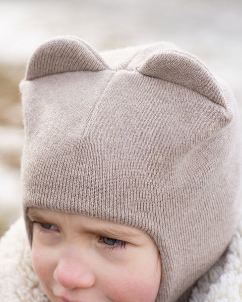 A close-up of a child's head, partially obscured, wearing a creamy beige menique knit balaclava or hat with two ear-like protrusions on top. The child's right eye and part of their face are visible, and they appear to be looking slightly to their right. A textured, light-colored collar or hood of a jacket is visible around their neck, and the background is blurred with light tones, suggesting an outdoor setting.
