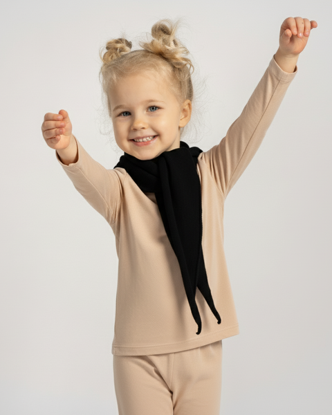 Happy child in a beige outfit with a black knit triangle scarf around the neck, arms raised playfully while smiling.