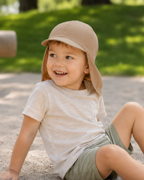 A smiling toddler sitting in a sandy play area outdoors, looking off to the side. He is wearing a beige short-sleeve t-shirt, olive green shorts, and a beige sun cap with a front brim and a protective neck flap.