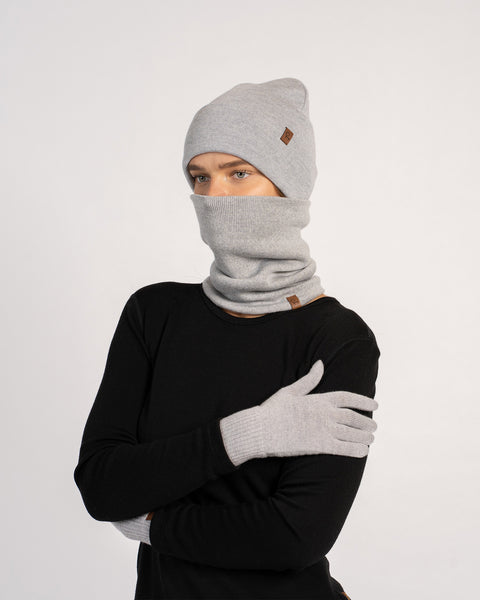 Vrouw draagt een lichtgrijze gebreide muts, bijpassende wanten en een nekwarmer die haar gezicht bedekt, gekleed in een zwart shirt met lange mouwen, poseert met gekruiste armen tegen een effen achtergrond.