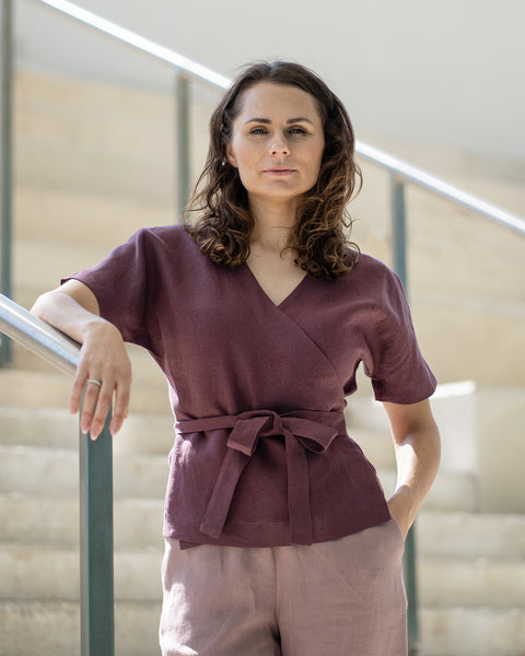 Een vrouw met bruin haar, gekleed in een donkerpaarse wikkelblouse en licht mauve broek, glimlacht terwijl ze poseert op betonnen binnentrappen.