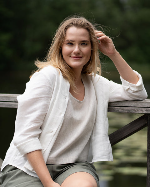 A woman sits casually on a wooden railing near a tranquil body of water, smiling softly at the camera. She is dressed in a relaxed white shirt layered over a light top and olive green shorts.