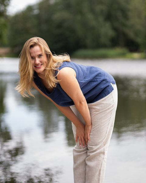 A woman stands outdoors with a river behind her, leaning forward cheerfully with her hands resting just above her knees. She is wearing a dark blue sleeveless linen top and loose-fitting natural beige linen trousers.