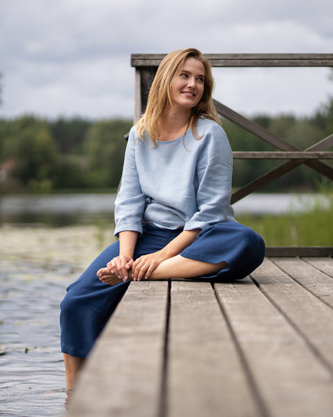Sitting cross-legged on a wooden dock next to a calm lake, a woman smiles brightly as she looks to the side. She is dressed comfortably in a light blue linen top and dark blue wide-leg pants.