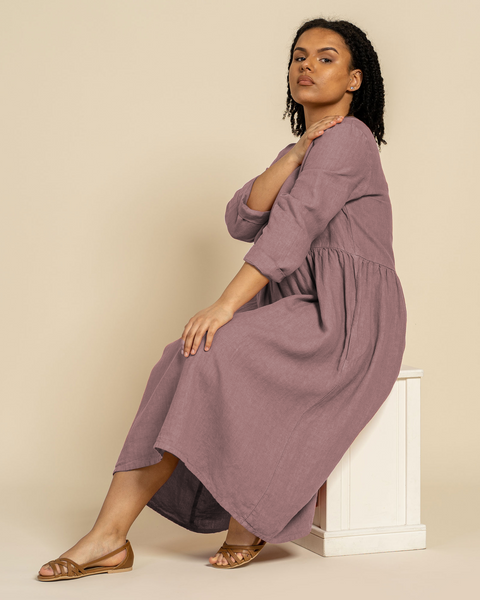 A woman with dark curly hair sits on a white block, looking towards the camera. She is wearing a long-sleeved menique linen dress and sandals. Her left hand rests on her right shoulder. The background is a solid light brown.