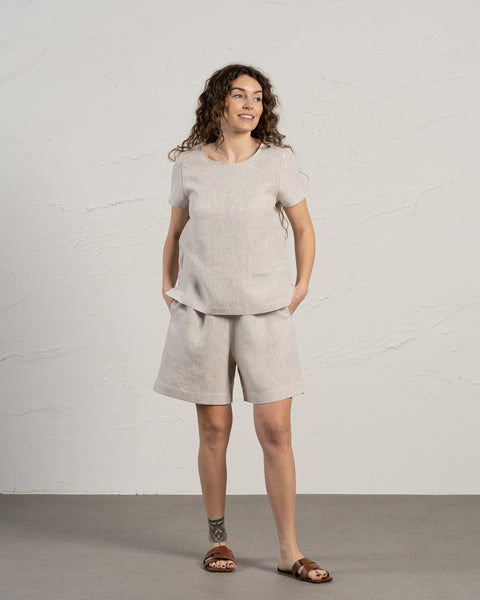 Een vrouw met bruin krullend haar draagt de menique bermudashort van 100% linnen en een linnen t-shirt top set. Haar handen zitten in de zakken terwijl ze zijwaarts kijkt. De achtergrond van de foto is licht en in koele tinten.
