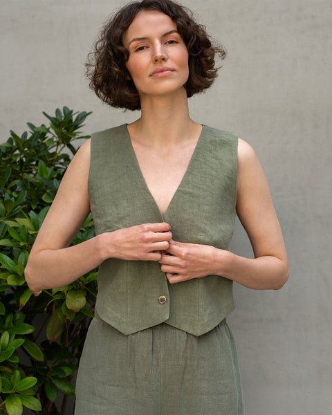 Een vrouw met kort, donker, krullend haar staat recht vooruit en kijkt iets omhoog. Ze draagt een mouwloos, steengroen linnen vest. Haar handen zijn bij de knoop, alsof ze die aanpast of vastmaakt. Ze draagt ook bijpassende steengroene linnen broek.