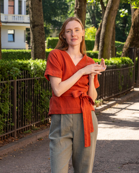 Een persoon met een lichte huid en donker haar staat in profiel, kijkt naar beneden naar hun handen die gekruist zijn ter hoogte van de buik. Ze dragen een linnen wikkelshirt met korte, losse mouwen en brede broekspijpen in steengroene kleur. Op de achtergrond is een donker metalen hek en weelderig groen gebladerte te zien.