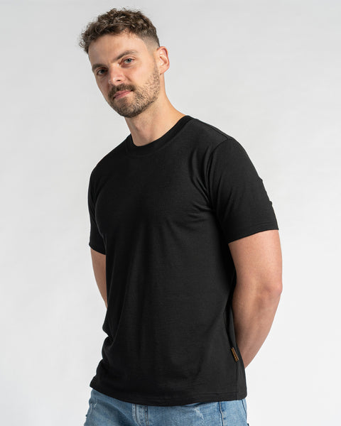 Een man die een zwart t-shirt van merinowol en een blauwe spijkerbroek draagt, is van borsthoogte te zien tegen een lichte achtergrond. Hij heeft kort bruin haar en een lichte baard en kijkt iets opzij. Het zwarte t-shirt van menique staat centraal en toont de pasvorm en het uiterlijk.