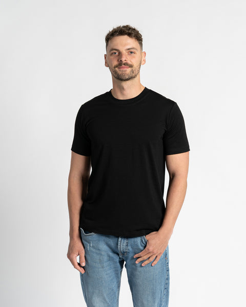 Een man met kort, donkerbruin haar en een keurig getrimde baard staat in het midden tegen een effen witte achtergrond, recht naar de kijker gericht. Hij draagt een zwart T-shirt met ronde hals en korte mouwen van het merk menique, dat de pasvorm en eenvoud van het kledingstuk laat zien. Het T-shirt is gemaakt van merinowol. Hij draagt ook lichtblauwe spijkerbroeken met een rechte pijp en een zichtbaar voorvak, waarbij hij één hand nonchalant in het vak steekt.
