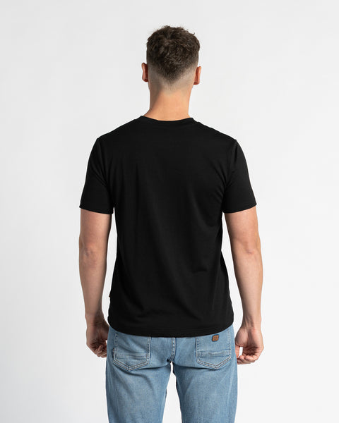 Achteraanzicht van een man die een zwart T-shirt en lichtblauwe spijkerbroek draagt, staand tegen een witte achtergrond. Het zwarte T-shirt staat centraal en toont de normale pasvorm en het ontwerp van achteren. De man staat in het midden, waardoor de achterkant van het kledingstuk duidelijk zichtbaar is.