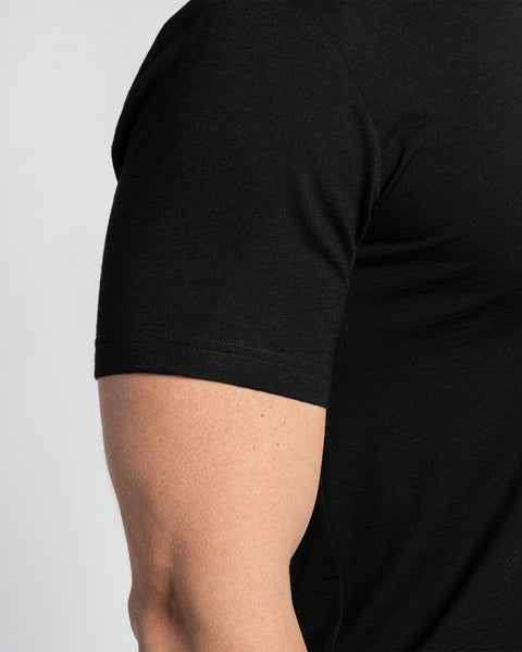 Close-up van een zwarte merino T-shirtmouw, met zicht op de stofstructuur en de naaddetails. Het T-shirt wordt gedragen aan een arm, met een lichte achtergrond.