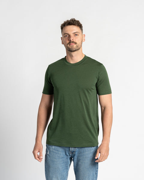 Een man draagt een donkergroen merinowollen T-shirt met ronde hals tegen een witte achtergrond. Het T-shirt staat centraal in de afbeelding en toont de kleur en pasvorm. Hij draagt ook lichtblauwe spijkerbroeken.