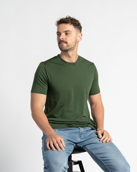 Man in een donker groen menique T-shirt en lichte spijkerbroek zit op een zwarte kruk, kijkt iets opzij, met een witte achtergrond.