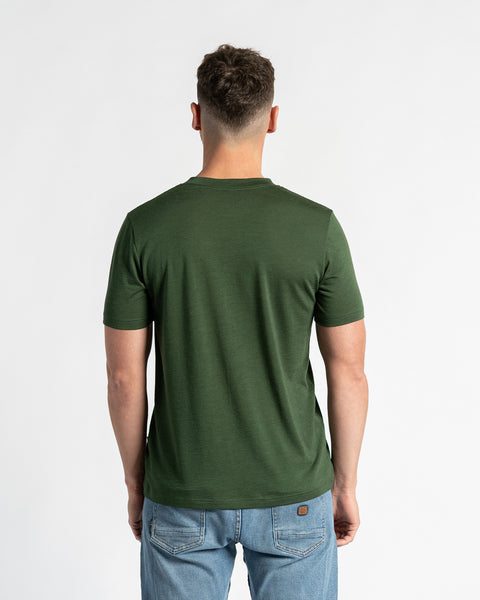 Achteraanzicht van een man in een donkergroen t-shirt van merinowol en lichtblauwe spijkerbroek, staand tegen een witte achtergrond.