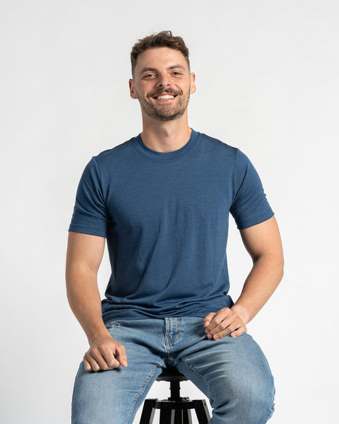 Een man draagt een t-shirt in spijkerstofkleur en lichtblauwe spijkerbroek en zit op een kruk tegen een lichte achtergrond. Hij heeft kort bruin haar, een lichte baard en lacht. Het menique t-shirt van merinowol staat centraal en toont de pasvorm en het uiterlijk terwijl de man zit.