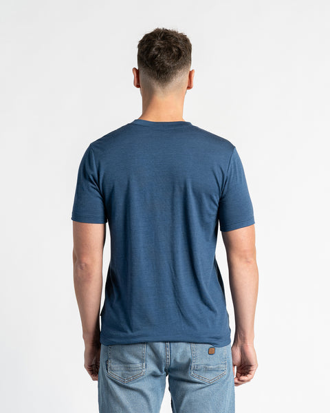 Achteraanzicht van een man die een menique t-shirt van merinowol en lichtblauwe spijkerbroek draagt, staand tegen een witte achtergrond. Het t-shirt in spijkerstofkleur is het belangrijkste aandachtspunt en toont de pasvorm en het ontwerp van achteren. De man staat centraal, waardoor de achterkant van het kledingstuk duidelijk zichtbaar is.