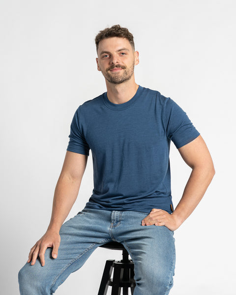 Een man draagt een t-shirt in spijkerstofkleur en lichtblauwe spijkerbroek en zit op een kruk tegen een lichte achtergrond. Hij heeft kort bruin haar, een lichte baard en glimlacht terwijl zijn hand op zijn heup rust. Het merinowollen t-shirt staat centraal en toont de pasvorm en het uiterlijk terwijl de man zit.