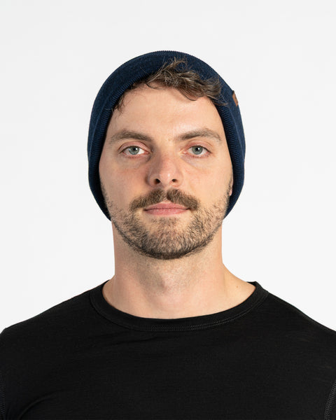 A person with light skin, short brown hair, a mustache, and light-colored eyes is shown in a headshot against a white background. They are wearing a blue knit beanie with a small brown tag on the side and a black long-sleeved shirt. They are looking directly at the camera.