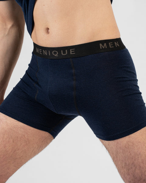 A close-up shot of a man's torso and thighs wearing navy blue short boxer briefs with a black "MENIQUE" waistband. He is lifting his matching navy t-shirt to reveal his midriff against a light grey background.