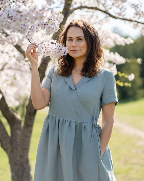 A woman with brown hair, wearing a light blue, short-sleeved linen wrap dress, stands outdoors next to a tree with white blossoms.