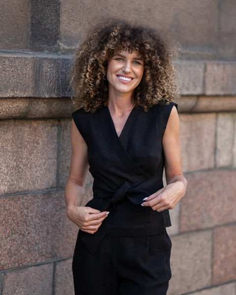 A smiling woman with voluminous, light brown curly hair stands outdoors, looking directly at the viewer. She is wearing a sleeveless black wrap top with a V-neck and a tied front, paired with menique linen black pants, though only the top portion is visible. Her hands are near the tie of her top.