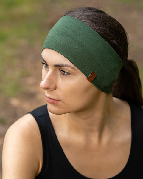 De foto toont een vrouw die in een bos zit, gekleed in de uniseks donkergroene menique merino HOEDEBAND. Ze draagt ook een mouwloos zwart merino hemd. Ze kijkt naar de zijkant.