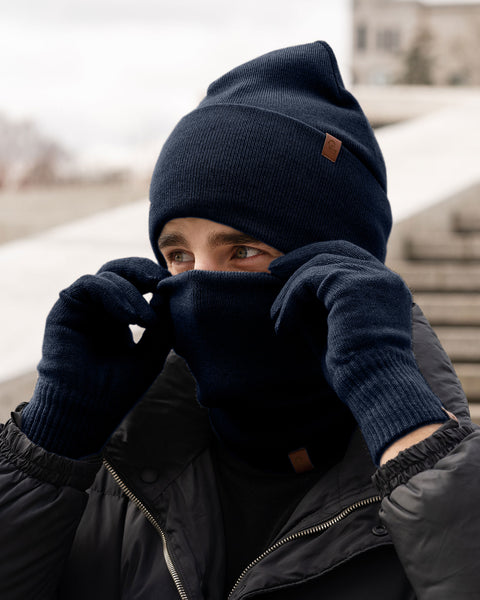 A person bundled in a dark blue menique knitted hat, a matching neck gaiter pulled up over their nose, and coordinating gloves. They are looking to their right, with their hands near their face as if adjusting their neck gaiter. A small brown tag is visible on both the hat and the neck gaiter. They are wearing a dark, possibly black, puffer jacket. In the background, there are blurred concrete steps and what appears to be an outdoor urban setting.