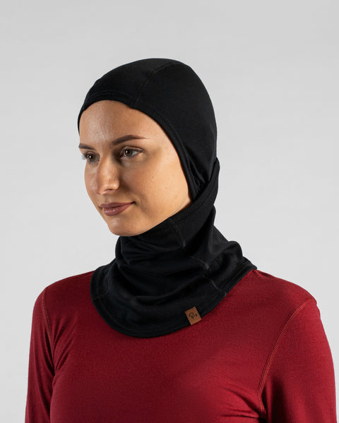 A studio portrait of a blonde woman looking off to the left, wearing a burgundy long-sleeved shirt and a black balaclava pulled up over her head, with the face section pulled down under her chin to leave her face completely exposed.