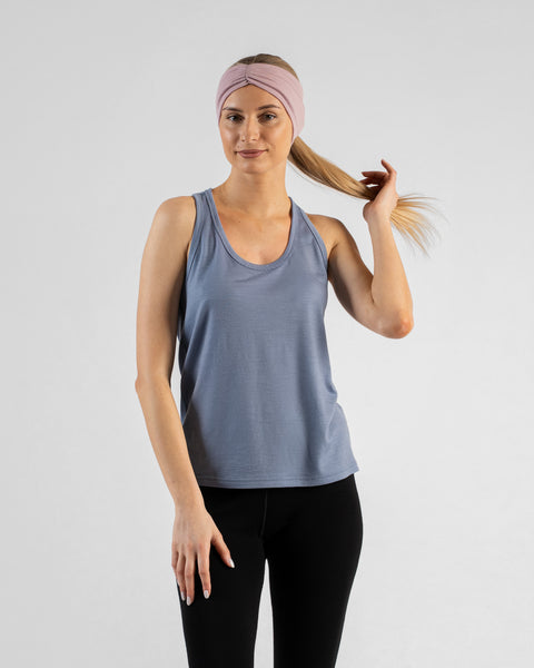 A studio portrait of a blonde woman standing and looking directly at the camera with a subtle smile against a plain light grey background. She is wearing a dusty blue sleeveless top, black leggings, and a dusty pink ribbon headband, with one hand casually holding the end of her ponytail.