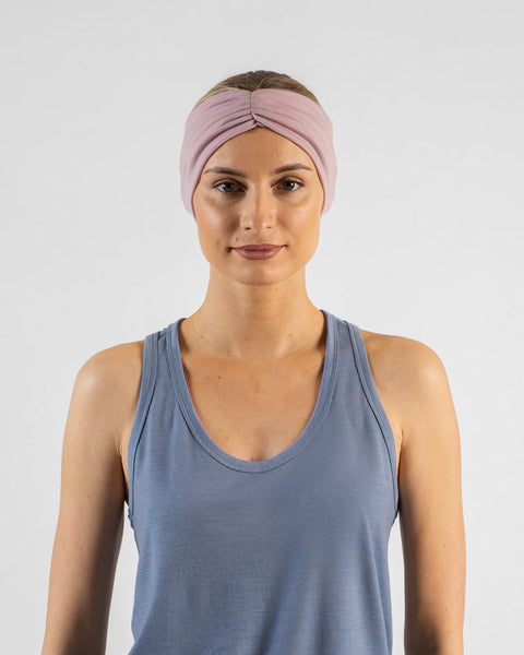 A studio portrait of a blonde woman facing forward and looking directly at the camera against a plain light grey background. She is wearing a dusty blue sleeveless top and a dusty pink ribbon headband.
