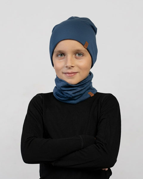 Een kind draagt een donkerblauwe muts en een nekwarmer set met een zwart shirt met lange mouwen, poseert met gekruiste armen en een lichte glimlach tegen een lichtgrijze achtergrond.