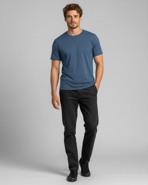 Een man loopt vooruit en draagt een blauw T-shirt, zwarte spijkerbroek en zwarte sportschoenen, staand tegen een effen grijze achtergrond.