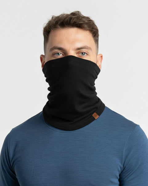 Man draagt een zwarte merinowollen nekwarmer met een blauw ondershirt tegen een lichte achtergrond.
