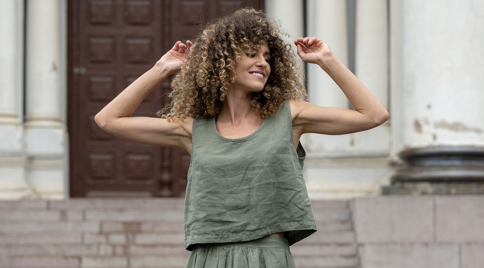 a woman standing by a building, wearing a full black linen set, that includes black pants and a black linen button-up shirt. She is smiling and has curly hair.