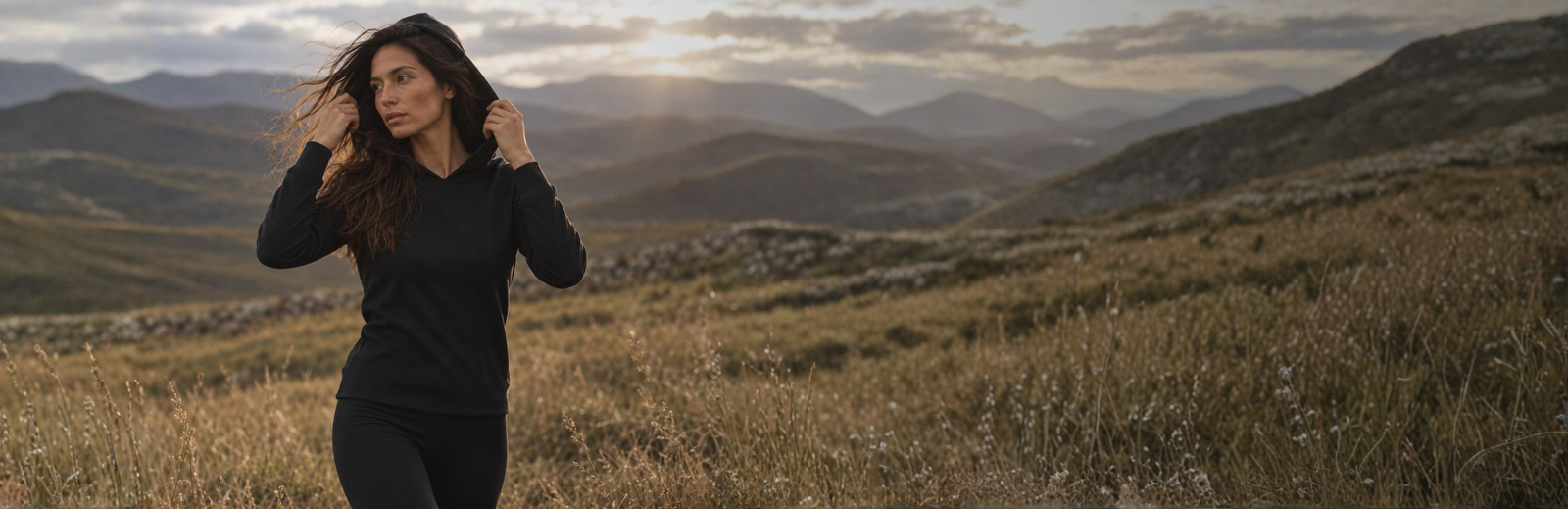 A woman wearing a black hoodie stands in a field of dry grass, holding the edges of her hood as the wind blows her hair. The sun shines through a cloudy sky over the rolling mountains behind her.