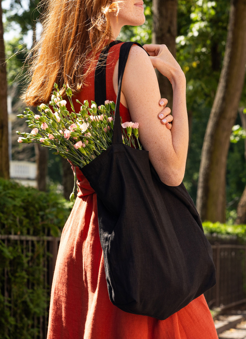 A woman wearing a sleeveless rust-colored linen dress is standing outdoors in a sunlit park, carrying a large black fabric tote bag filled with fresh pink and white flowers over her shoulder. The scene feels calm and natural, with green trees in the background and soft sunlight highlighting the texture of the linen fabric and the bouquet.
