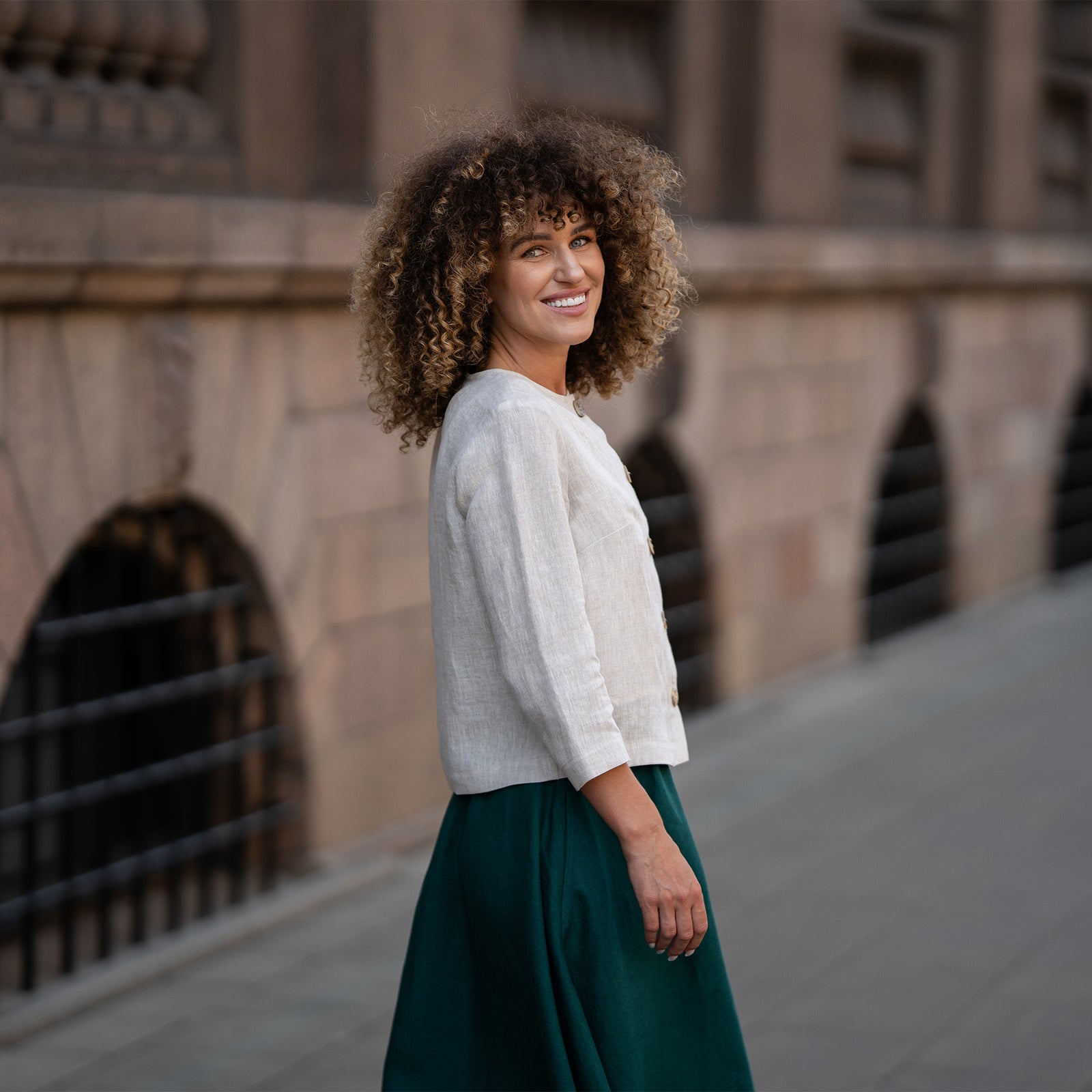Een model draagt een natuurlijk beige linnen jasje Alice met knopen, met een losse pasvorm, driekwart mouwen en een minimalistisch ontwerp. Het jasje is gecombineerd met een bijpassende donkergroene rok.
