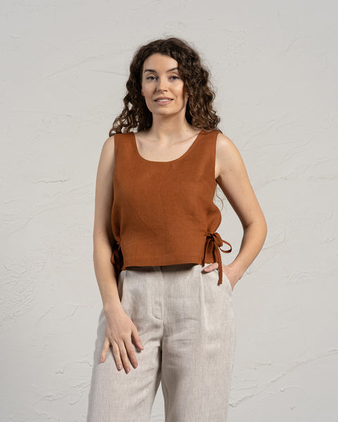 A smiling woman with long, curly brown hair wears a sleeveless linen crop top with side ties and light-colored wide-leg linen pants. She stands in front of a textured light gray wall, with one hand in her pocket.