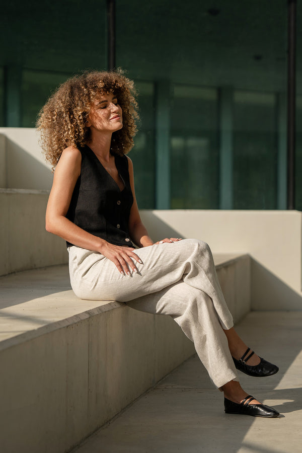 A woman with voluminous curly brown hair sits with her eyes closed on a set of concrete steps, seemingly enjoying the sunlight. She is wearing a dark menique sleeveless top, light-colored, loose-fitting linen trousers, and black ballet-style shoes with straps. The background features a modern building with large, dark windows, and the shadows suggest it is a bright day.