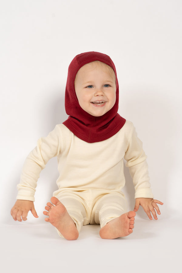 Een peutermeisje zit op de grond, draagt de menique kinder merino 160gsm bivakmuts in koninklijk kersenkleur, studioachtergrondfoto.