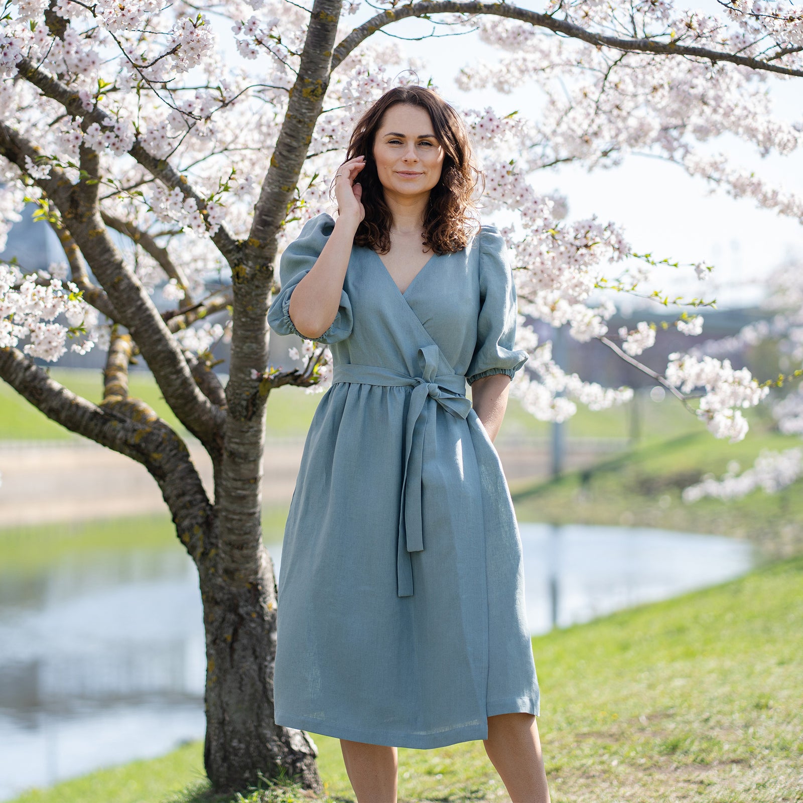 A light-skinned person with wavy brown hair has their eyes closed and appears to be smelling a cluster of white blossoms held in their right hand. They are wearing a linen menique wrap dress with short puff sleeves and a tied waist, and their left hand is in a pocket. A blossoming tree and a blurred green background suggest an outdoor setting.